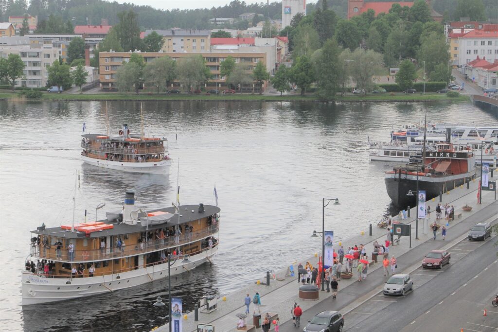 Kaupungin satamaa, jossa kolme vanhaa laivaa näkyvissä sekä ihmisiä kadulla.