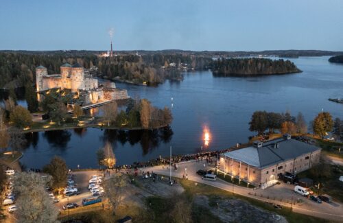 Ilmakuva Olavinlinnasta ja tiilisestä museorakennuksesta sekä iltahämärässä oleva järvimaisema. Järvessä palaa kokko, ja ihmisiä on paljon rannalla.