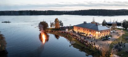 Ilmakuvassa tiilinen museorakennus, järvessä palava kokko, ja rannalla paljon ihmisiä iltahämärässä.