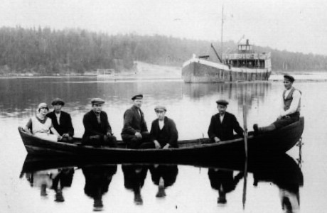 Vanhassa kuvassa ihmisiä istuu soutuveneessä, jonka takana on iso laiva.