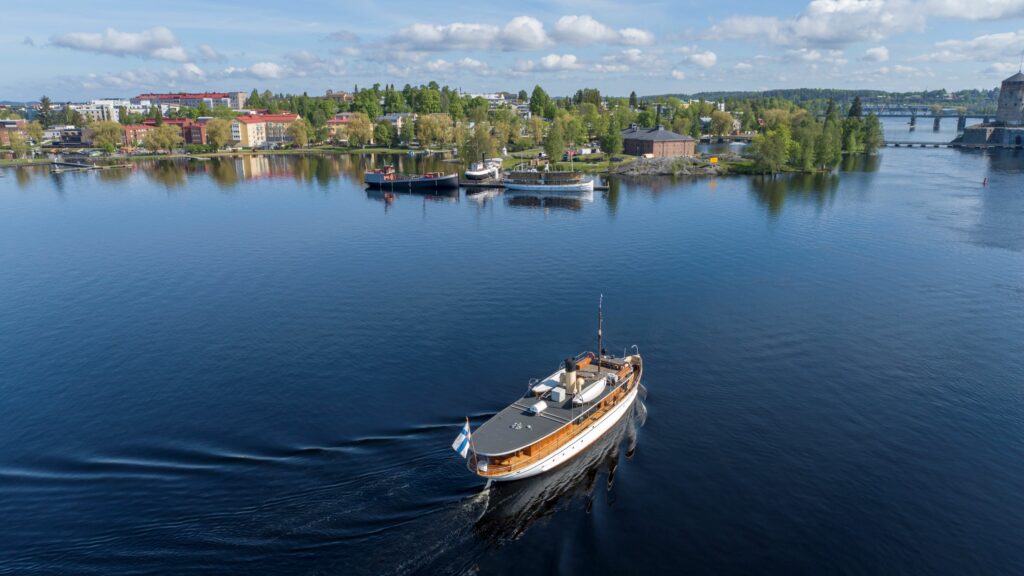 Vanha laiva seilaa kohti laivalaituria, jossa kolme muuta museolaivaa. taustalla museorakennus sekä Olavinlinna.
