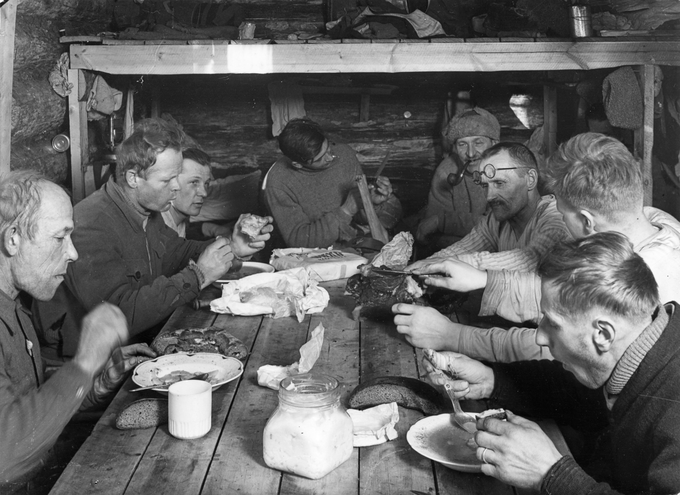 Vanhassa kuvassa miehiä syömässä lankkupöydän ääressä.