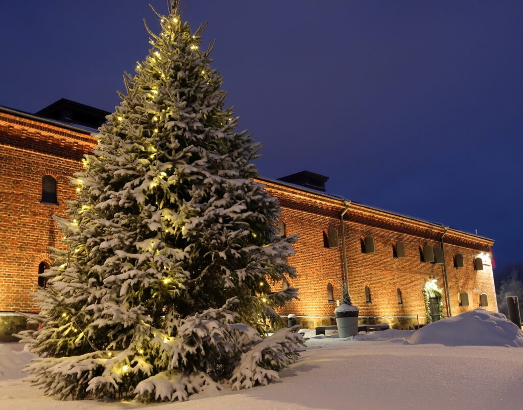 Joulukuusi valaistun tiilisen museorakennuksen edessä. Hämärä iltataivas taustalla sekä lumihankea.