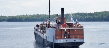 Vanha puna-harmaa-musta laiva kuvattu perästä päin. Seilaa kesäkelissä järvellä ja kyydissä on ihmisiä.