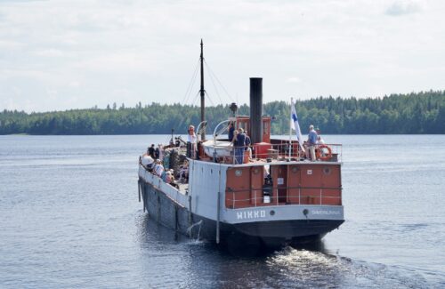 Vanha puna-harmaa-musta laiva kuvattu perästä päin. Seilaa kesäkelissä järvellä ja kyydissä on ihmisiä.