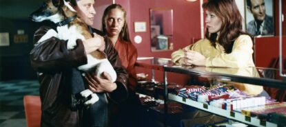 Kolme henkilöä, yhdellä pieni koira sylissä. Yksi henkilöistä nojaa myyntitiskiin, jossa makeisia esillä. Henkilöiden ilmeet vakavia.