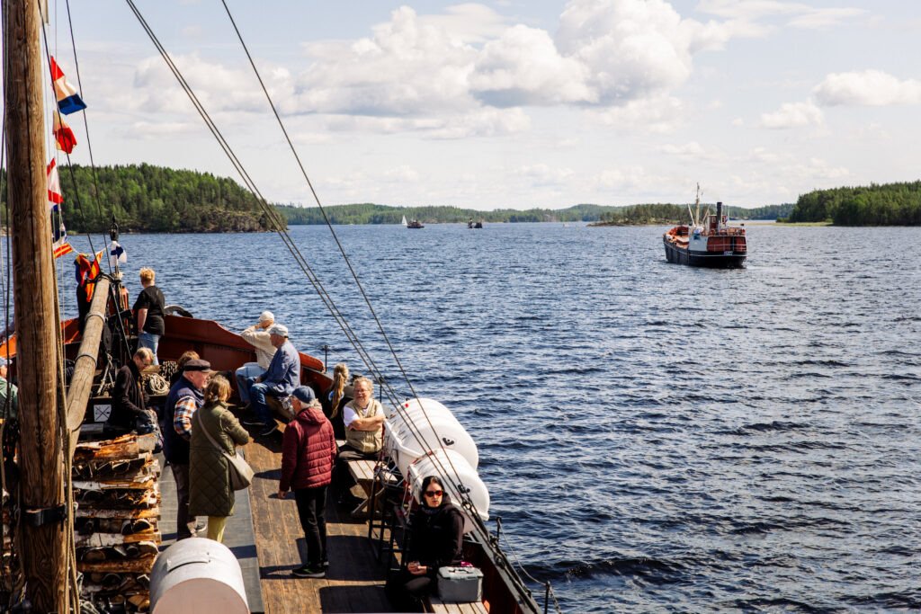 Sinisellä kesäisellä järvellä seilaa taustalla vanhoja laivoja. Etuvasemmalla vanha tervahöyry, jonka avokannelle ihmisiä kyydissä.