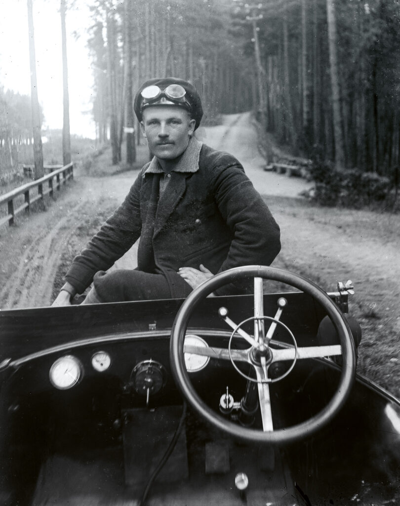 Miesoletettu istuu vanhanmallisen avoauton keulalla. Hänellä on ajolasit ja lakki päässä. Taustalla vanhaa hiekkaista metsätietä rinteessä.