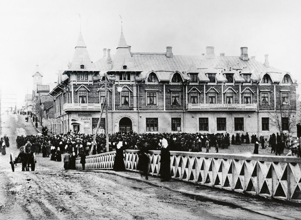 Taustalla vanha puinen kaksikerroksinen hotellirakennus, jossa suippoja torneja sekä erimuotoisia ikkunoita. Edessä sillankaide. Sillan ja rakennuksen välissä massoittain ihmisiä kokoontuneena tummissa vaatteissaan rakennuksen eteen. Myös hevoskärry näkyy.