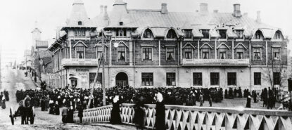 Taustalla vanha puinen kaksikerroksinen hotellirakennus, jossa suippoja torneja sekä erimuotoisia ikkunoita. Edessä sillankaide. Sillan ja rakennuksen välissä massoittain ihmisiä kokoontuneena tummissa vaatteissaan rakennuksen eteen. Myös hevoskärry näkyy.