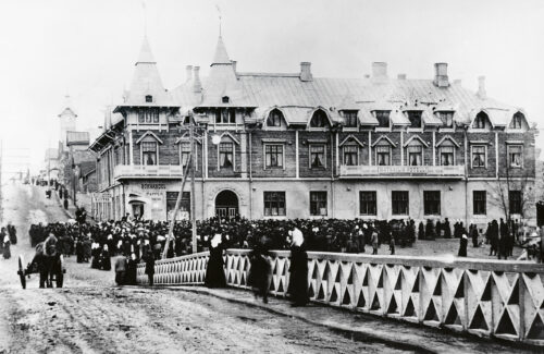 Taustalla vanha puinen kaksikerroksinen hotellirakennus, jossa suippoja torneja sekä erimuotoisia ikkunoita. Edessä sillankaide. Sillan ja rakennuksen välissä massoittain ihmisiä kokoontuneena tummissa vaatteissaan rakennuksen eteen. Myös hevoskärry näkyy.