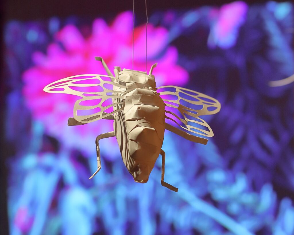 Valkoisesta paperista tehty lentävä hyönteinen. Taustalla violettia ja purppuraa.