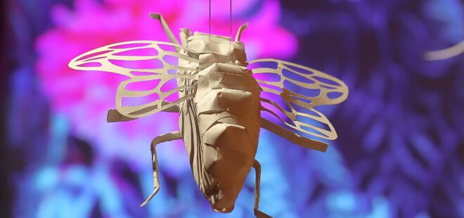 Valkoisesta paperista tehty lentävä hyönteinen. Taustalla violettia ja purppuraa.