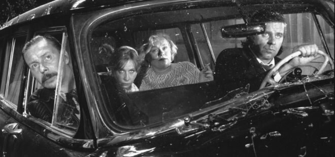 Neljä aikuista istuu vanhassa tummassa autossa. Kaksi naisoletettua on takapenkillä, ja toisella heistä on savuke kädessä.