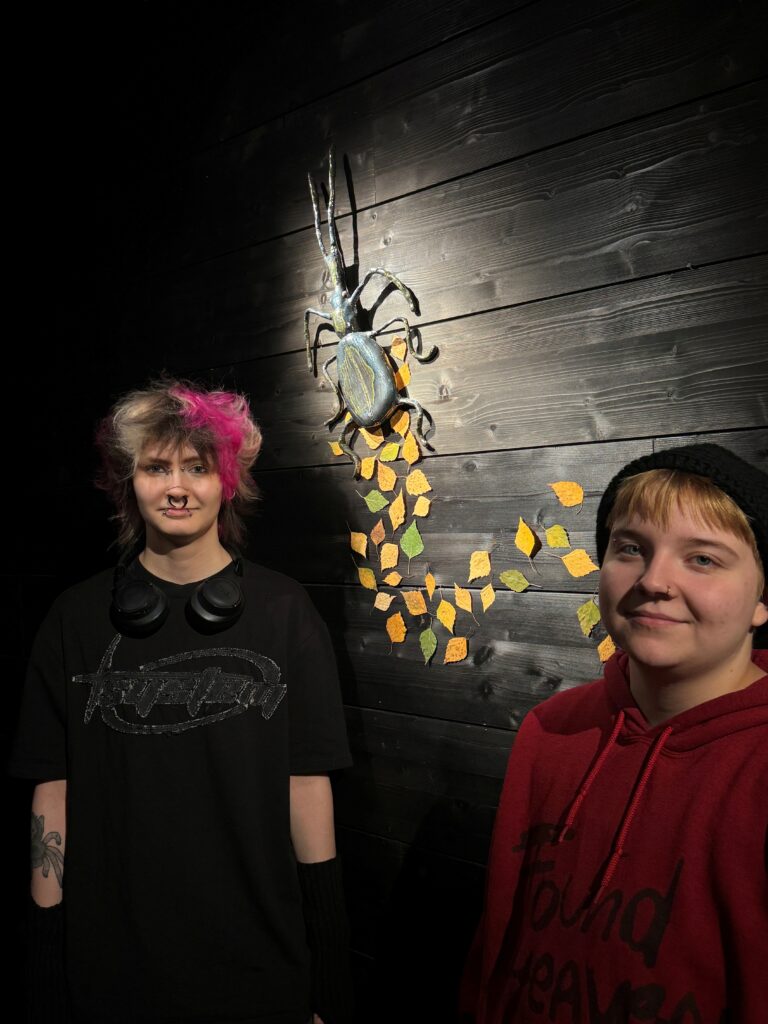 Pitkäsarvinen ja iso tumma kovakuoriainen mustalla seinällä. Alla keltaisia koivunlehtiä. Kaksi ihmistä seisoo teoksen edessä.