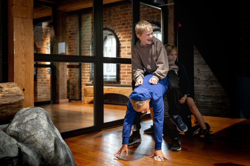 Kolme lasta touhuaa lattialla: yksi on kontillaan lattialla, ja kaksi muuta on hänen selässään. Lapset nauravat iloisesti.