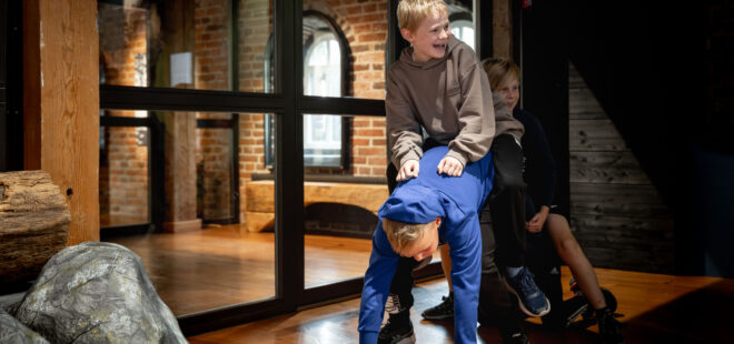 Kolme lasta touhuaa lattialla: yksi on kontillaan lattialla, ja kaksi muuta on hänen selässään. Lapset nauravat iloisesti.