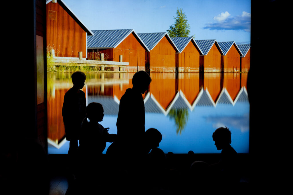 Seinällä iso värillinen kuva punaisista venevajoista eli vesirajassa olevista rakennuksista, joissa säilytettiin veneitä. Kuvan edessä lapsia, jotka näkyvät vain mustina silhuettihahmoina.