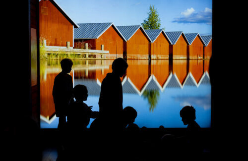 Seinällä iso värillinen kuva punaisista venevajoista eli vesirajassa olevista rakennuksista, joissa säilytettiin veneitä. Kuvan edessä lapsia, jotka näkyvät vain mustina silhuettihahmoina.