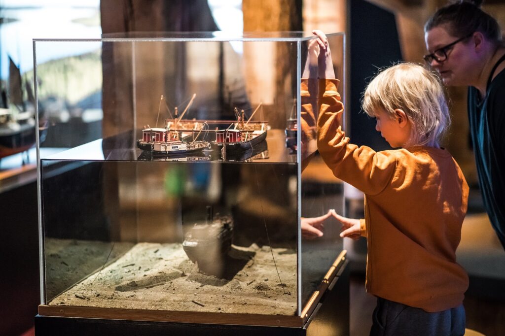 Lapsi ja aikuinen katsovat lasivitriiniä, jossa pienoismalleja vanhoista laivoista. Yksi laivoista näyttää olevan järven pohjassa, kun muut näyttävät olevan järven pinnalla.
