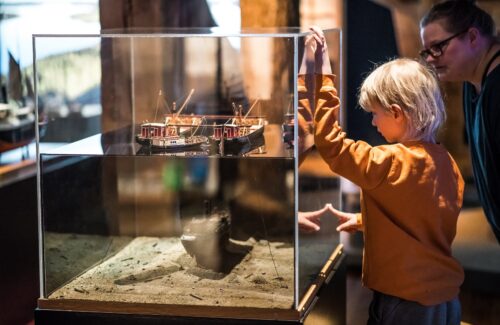Lapsi ja aikuinen katsovat lasivitriiniä, jossa pienoismalleja vanhoista laivoista. Yksi laivoista näyttää olevan järven pohjassa, kun muut näyttävät olevan järven pinnalla.