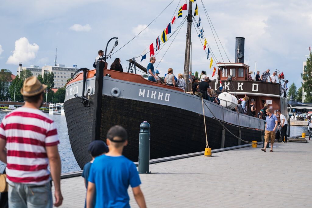 Vanha laiva puulaituriin kiinnittyneenä. Ihmisiä kannella kesävaatteissaan. Laivassa juhlaliputus sekä kyljessä teksti "Mikko".