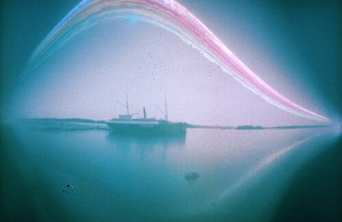 Utuinen värikuva, jonka keskellä vanha höyrylaiva laituriin kiinnittyneenä. Laivan yli kulkee sateenkaarimainen paksu juova, joka nousee keskellä laivan yläpuolella kaareksi.