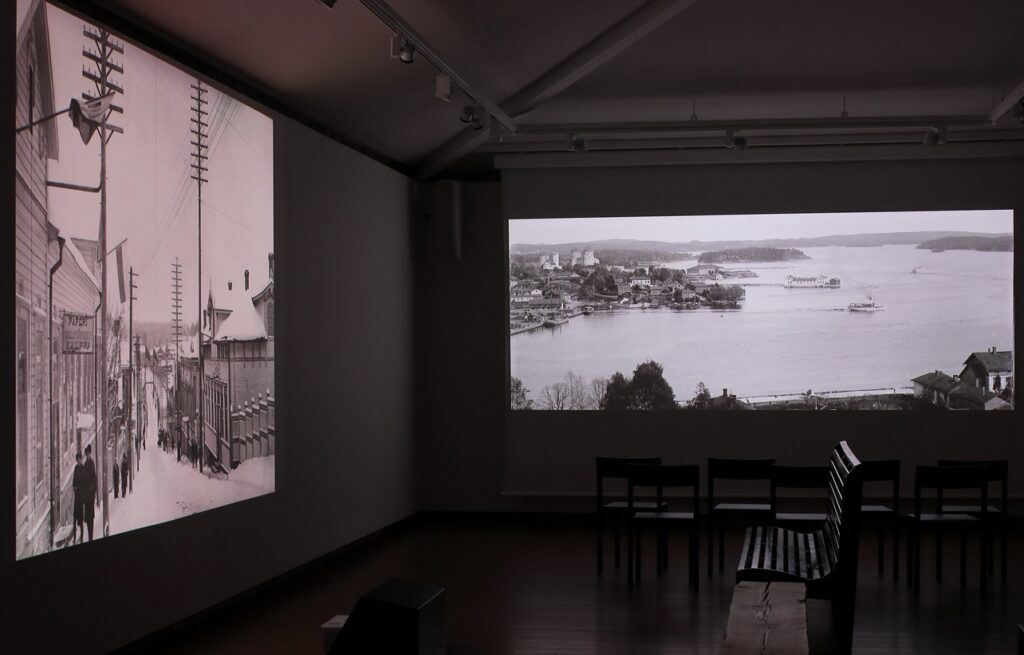 Hämärä huone, jossa kahdella seinällä suuret mustavalkoiset maisemavalokuvat.