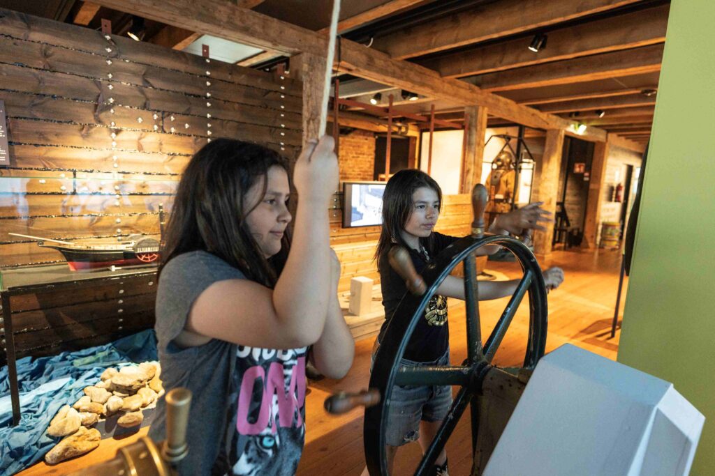 Kaksi pitkähiuksisista lasta. Toinen pyörittää suurta puista ruoria ja toinen vetää paksusta vanhasta köydestä.