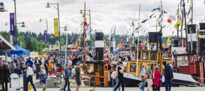 Iso tungos kaikenikäisiä ihmisiä, jotka katselevat satamalaiturissa kiinni olevia vanhoja värikkäitä höyrylaivoja.