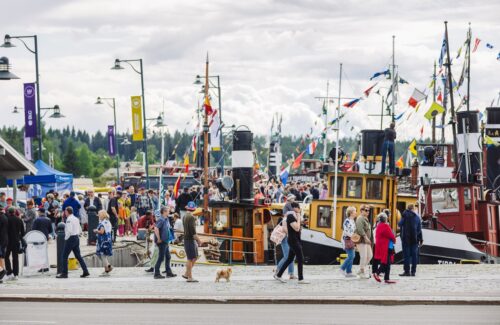 Iso tungos kaikenikäisiä ihmisiä, jotka katselevat satamalaiturissa kiinni olevia vanhoja värikkäitä höyrylaivoja.