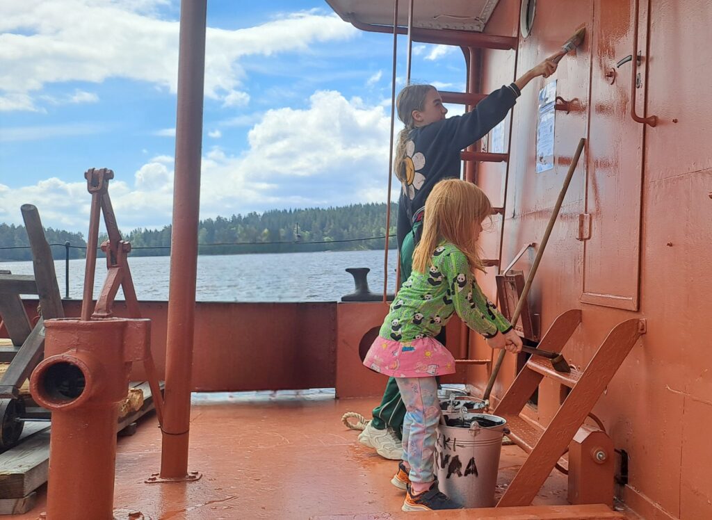 Kaksi lasta maalaa pensseleillä ja vedellä oranssinväristä laivan seinää. Vieressä peltinen sanko, jossa lukee "Leikkitervaa".