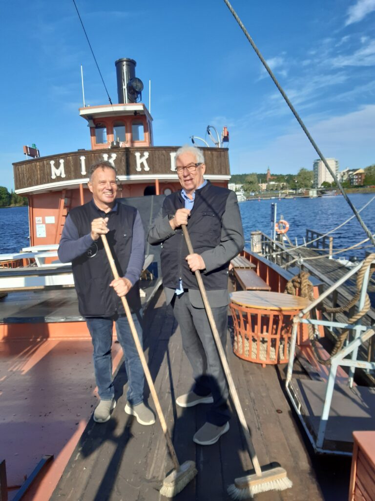 Kaksi miestä seisoo vanhan laivan kannella katuharjat kädessään.