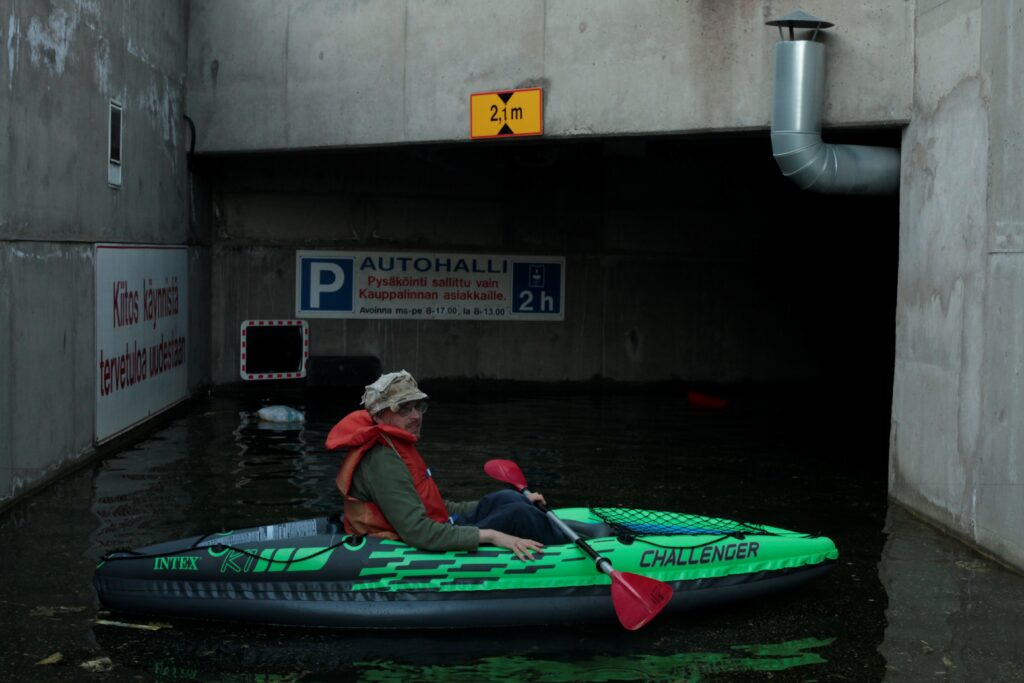 Aikuinen meloo ilmatäytteisellä vihreällä kajakilla vedentäyttämässä parkkihallin sisääntulorampissa.