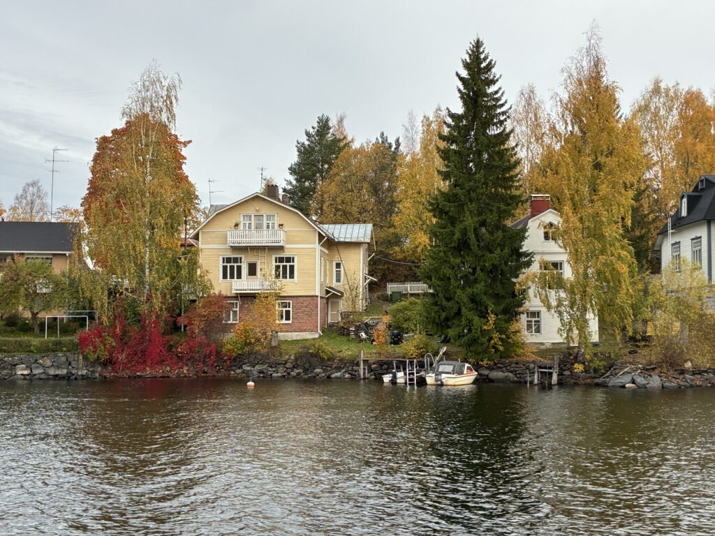 Kolme rakennusta pilkottaa syksyisten puiden takaa. Talot ovat järven rannalla, ja rannassa on laituri ja kaksi venettä. Kolme taloista on vanhoja puutaloja.