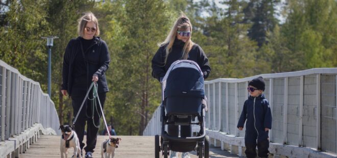 Kaksi naispuolista henkilöä ja pieni poika kävelysillalla. Toinen työntää lastenvaunuja ja toisella on kaksi pientä koiraa hihnassa.